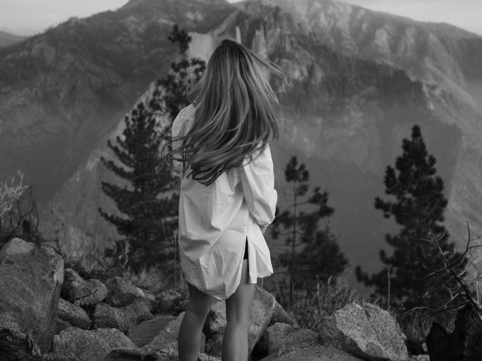 A woman in a white shirt with windblown hair looking out over a mountain valley